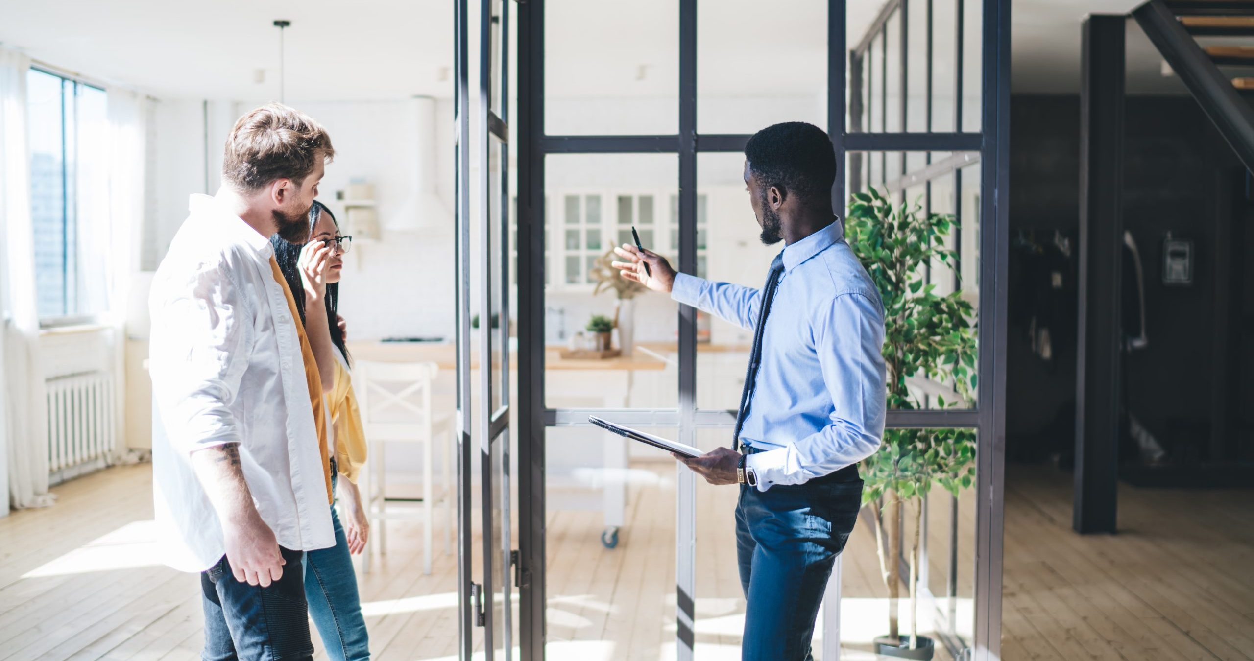 "A man gestures to show off the inside of a home, whilst he is stood inside with a couple"